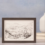 Framed print of a landscape drawing against a blue wall next to a small vase on books.