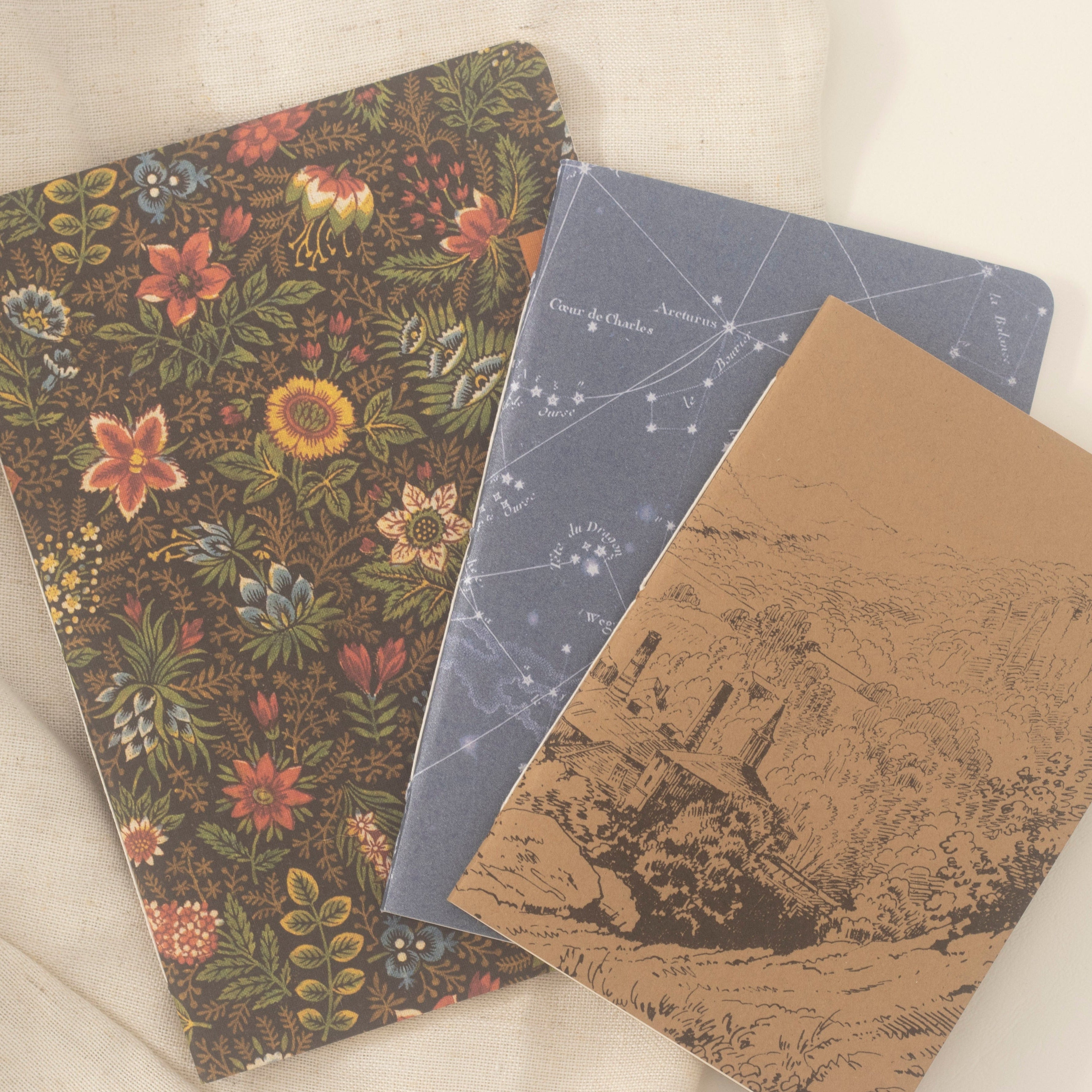 Three decorative notebooks arranged on a light beige fabric background. 
