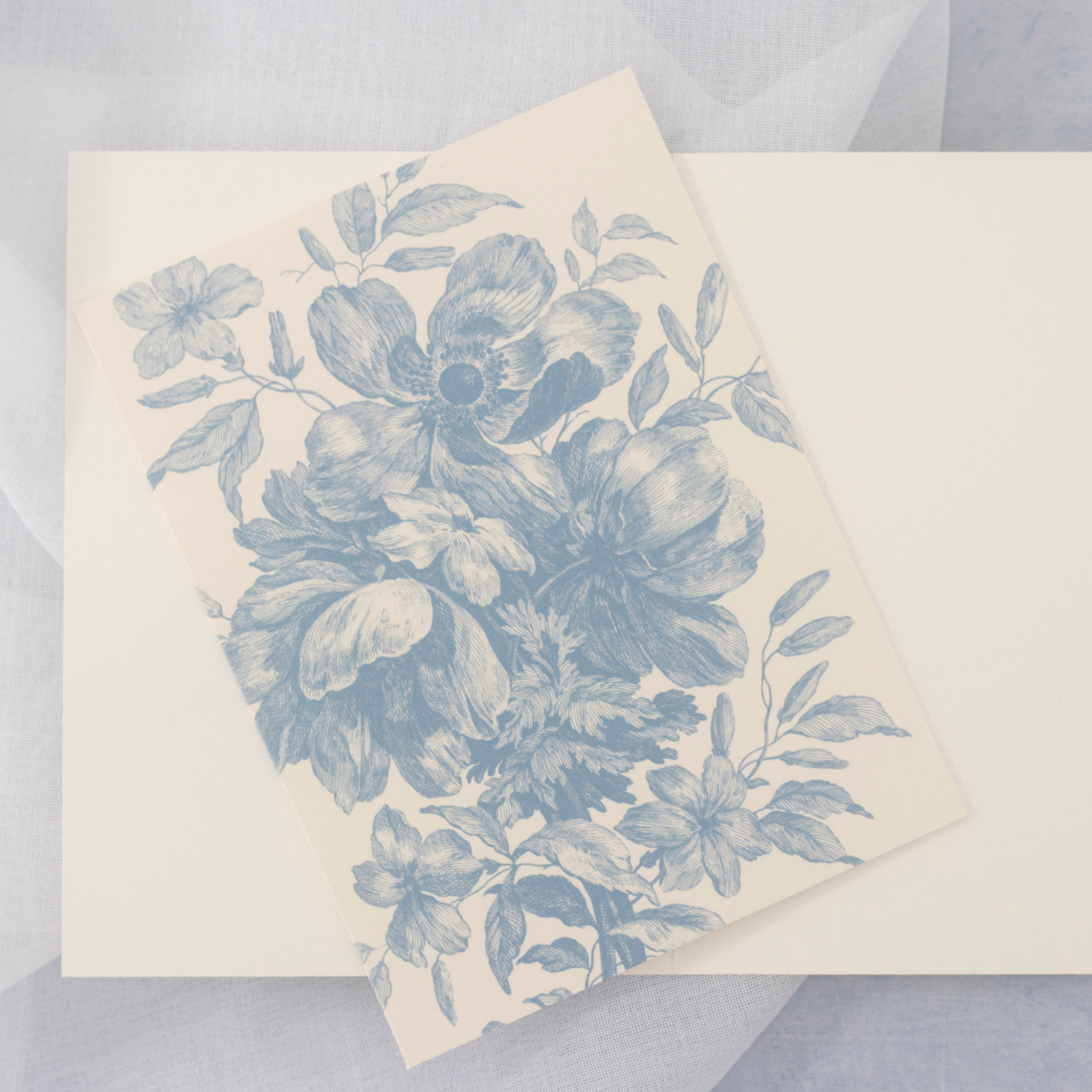 A cream-colored card featuring a detailed blue floral toile pattern, resting diagonally on an open card against a soft, light gray textured fabric background.