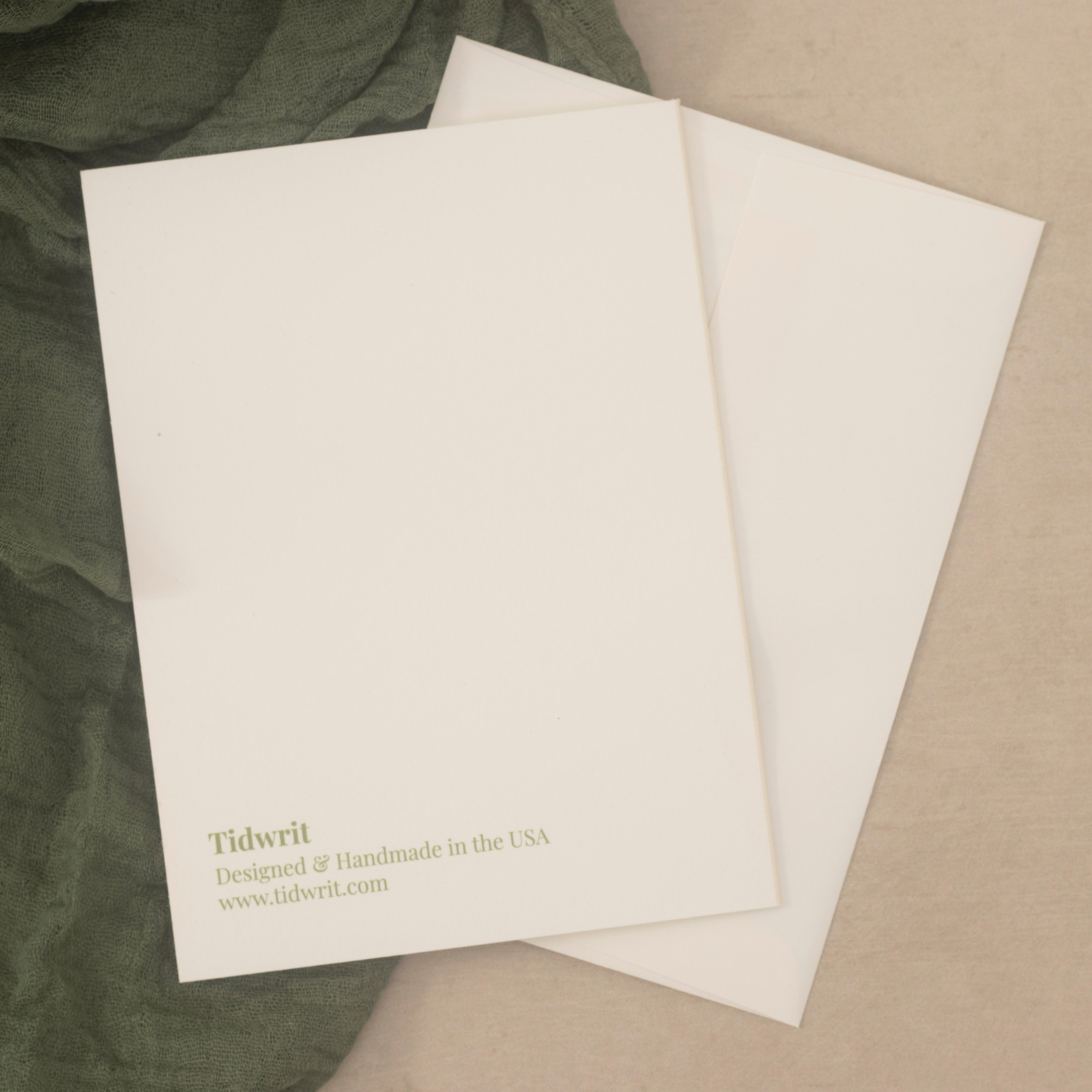 Back of an off-white stationery card on top of a matching off- white envelope over a textured dark green fabric and beige surface.