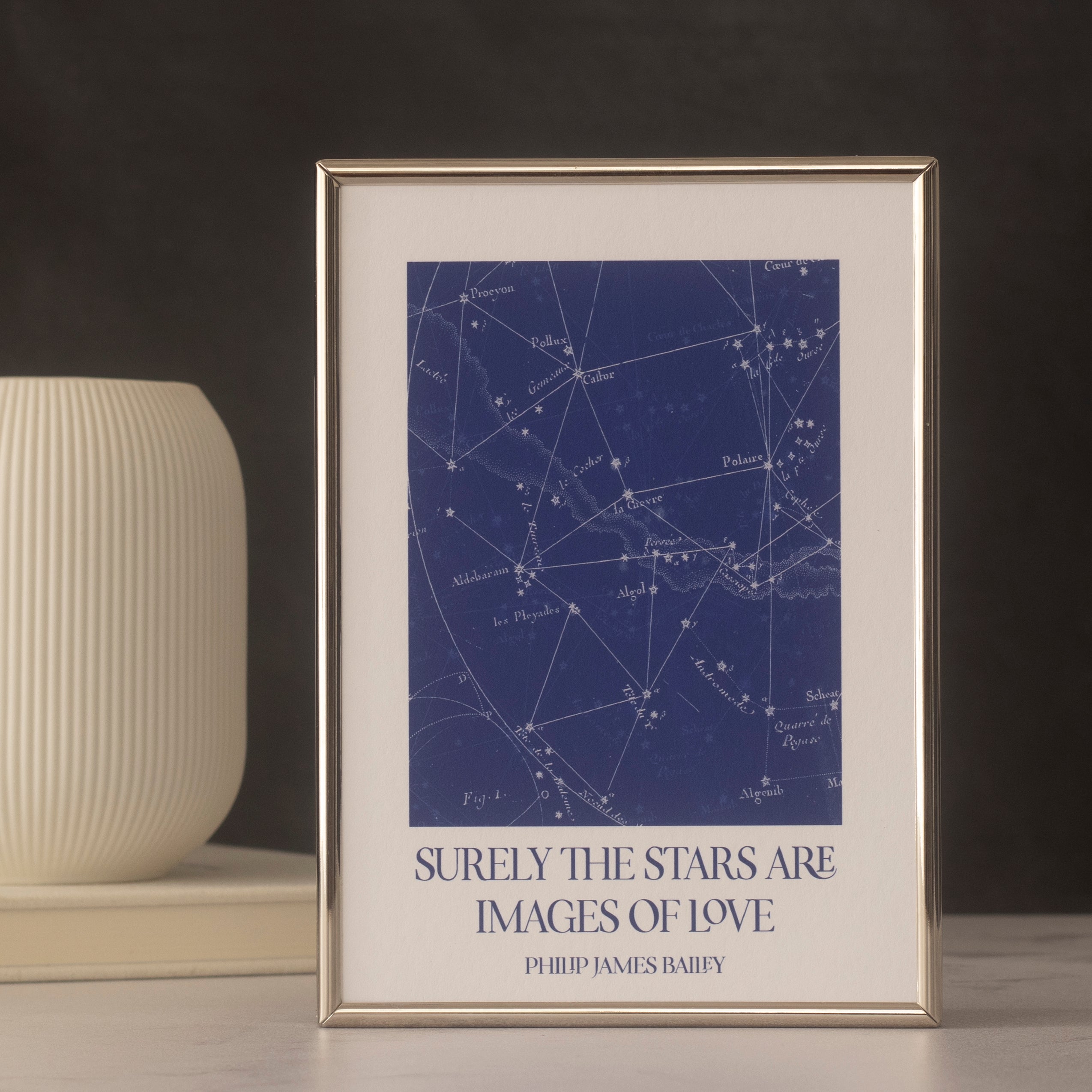 Framed star map print with a quote on a dark surface with a vase and book in the background.