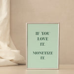 A framed typographic art print with a light green background and dark green text: 'IF YOU LOVE IT. MONETIZE IT. The background is a light beige with fabric draped on the left.