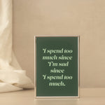 A print with a dark green background and light green text reading, 'I spend too much since I'm sad since I spend too much.' It has a thin metallic frame and is displayed standing on a tabletop against a warm beige wall, with soft draped fabric on the left side.