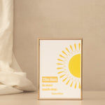 A gold-framed print featuring a yellow sun and the quote 'The Sun is new each day. ' and the text Heraclitus on a cream background, styled on a light beige background next to a draped fabric.