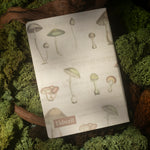 The back of a notebook with a vintage mushroom pattern on its cover is styled on a bed of green moss and wood.