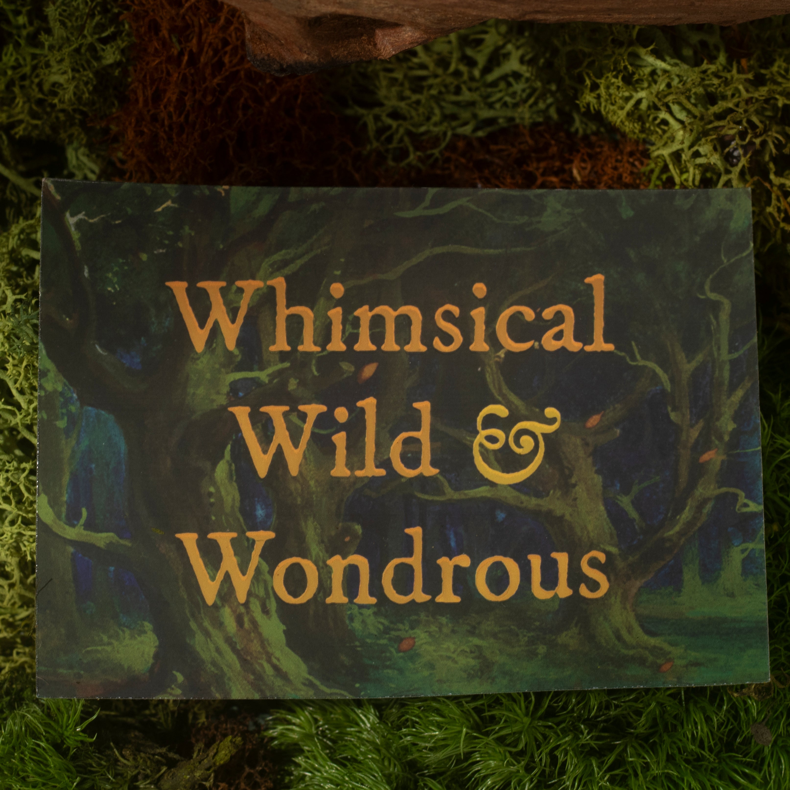A sticker with the phrase 'Whimsical Wild & Wondrous' in gold lettering over a dark forest illustration, placed on a bed of green moss.
