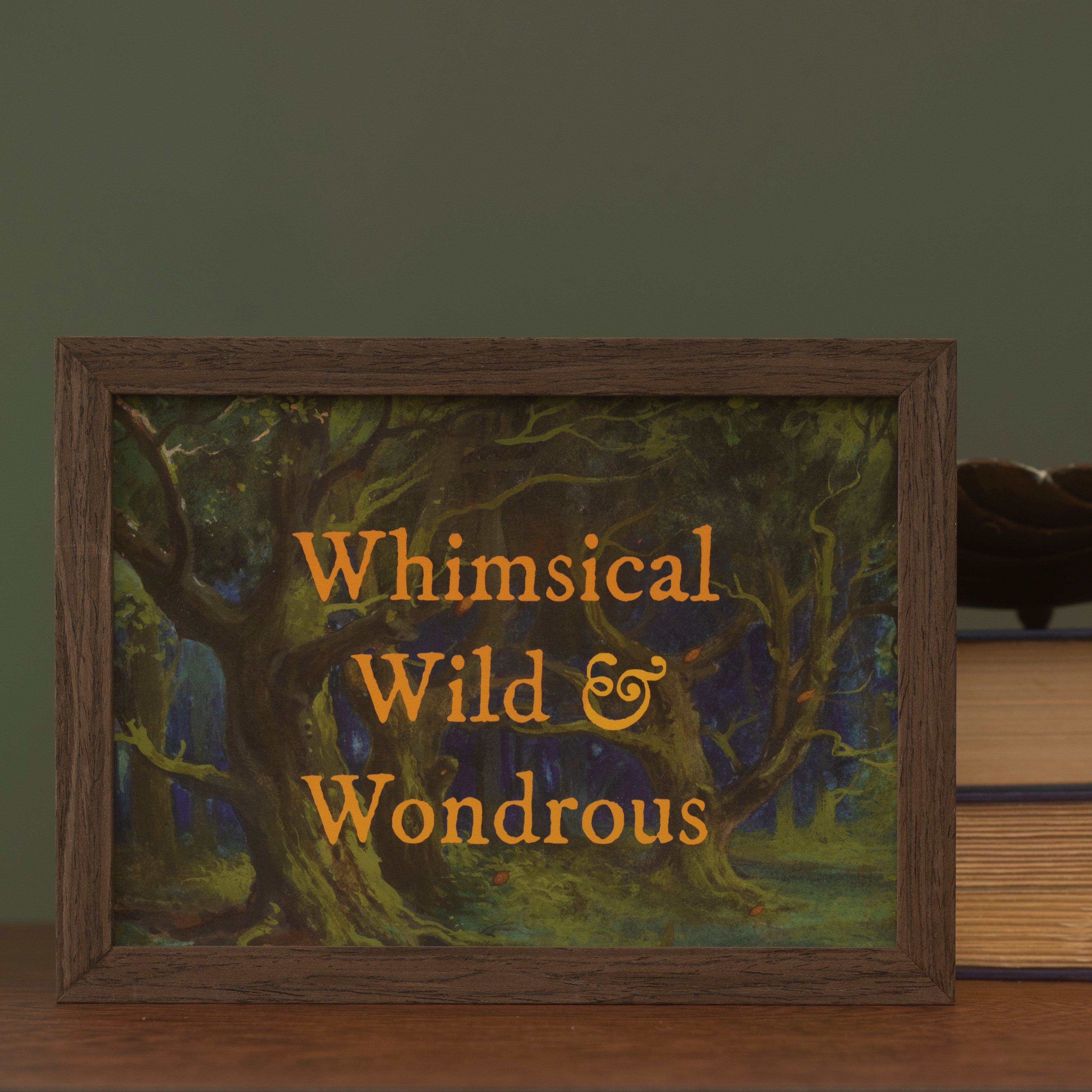 A framed art print featuring a dark forest scene with the text 'Whimsical Wild & Wondrous' in orange lettering, on a wooden surface against a dark green background.