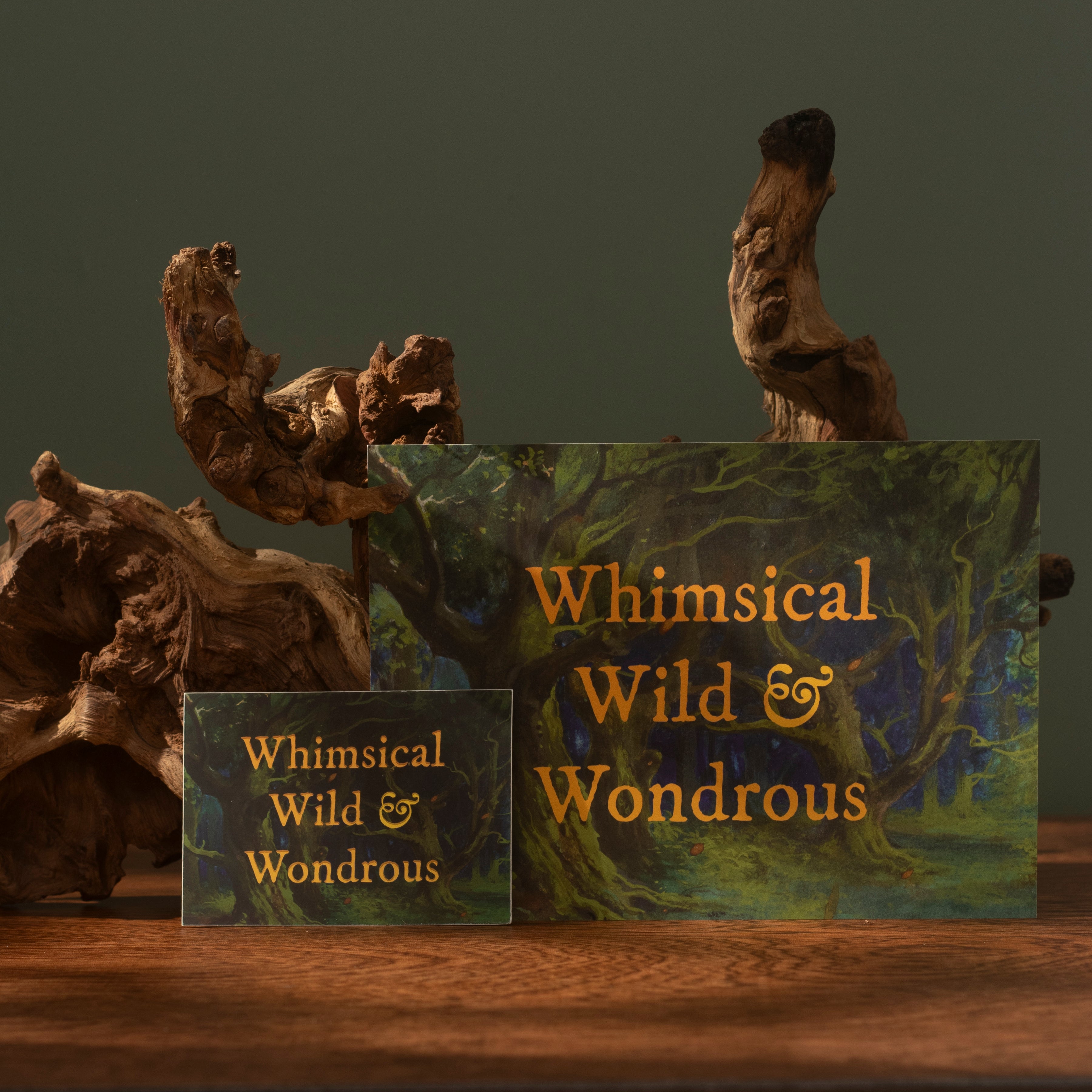 A sticker and a print with the phrase 'Whimsical Wild & Wondrous' in gold lettering, displayed on a wooden surface with driftwood props against a dark green background.