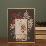 A framed botanical art print with a gold frame, displayed on a wooden surface next to books.