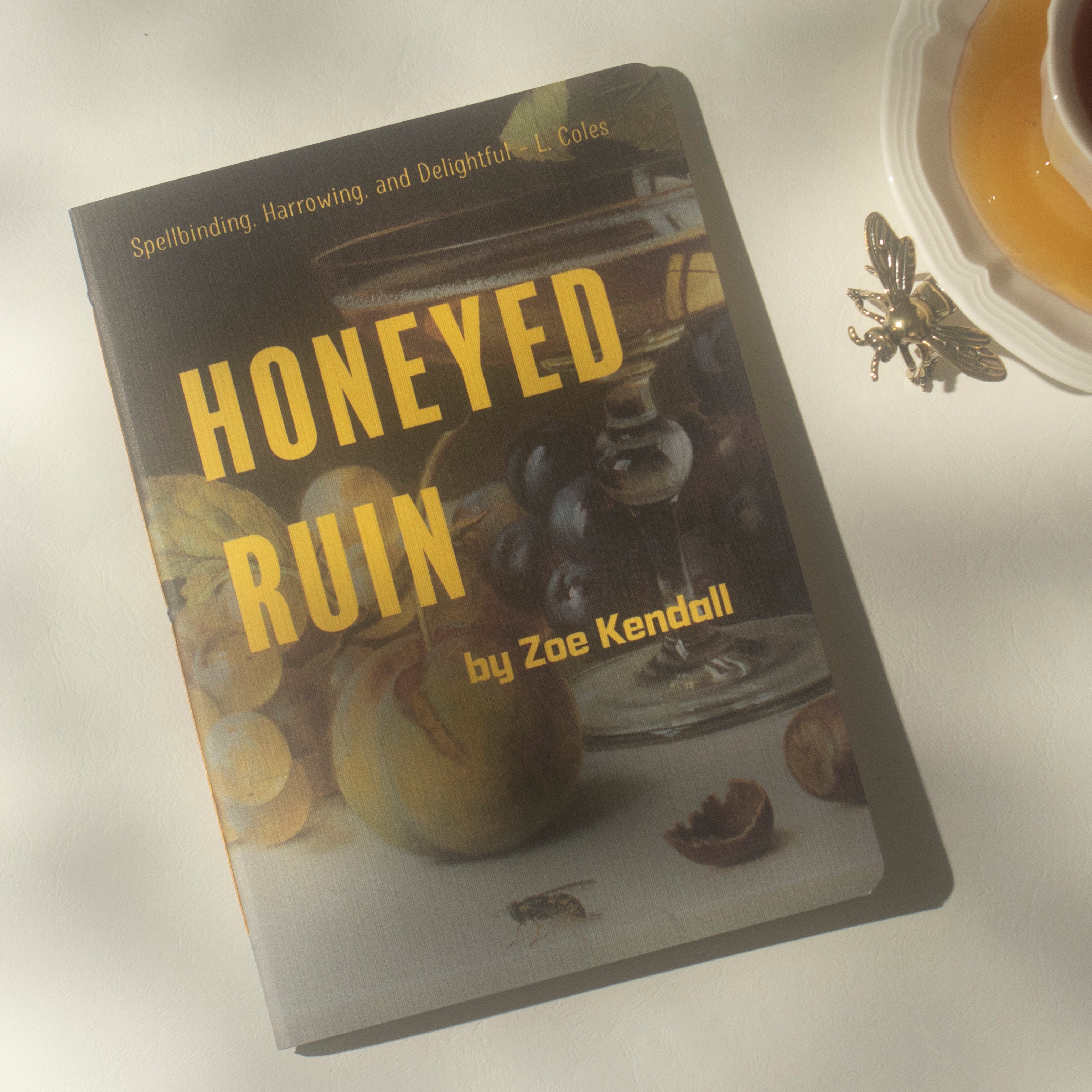 A notebook titled 'HONEYED RUIN' by Zoe Kendall, featuring a dark cover with large yellow title text over a still life photographed on a light surface next to a teacup and a decorative bee ornament.