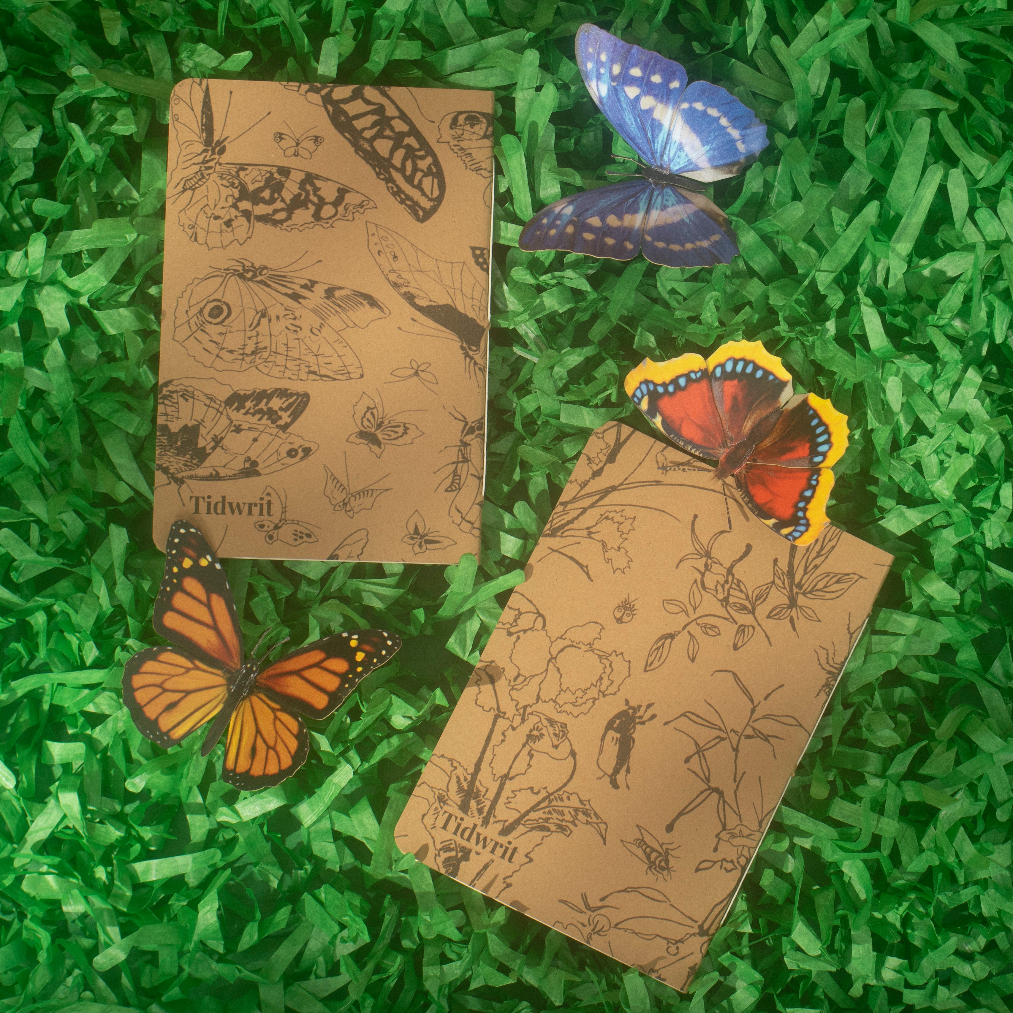 Backs of two kraft brown notebooks with insect, and floral illustrations on a paper grass background